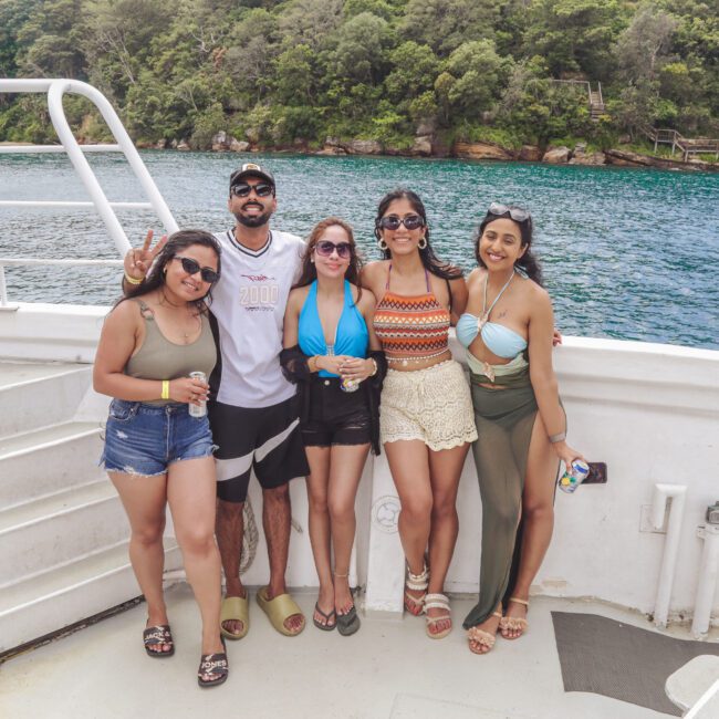 Five people stand together smiling on a boat deck, dressed in summer clothing and swimwear, with lush green trees and blue water visible in the background.