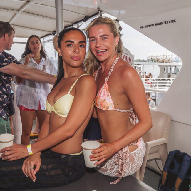 Two women in bikinis smile and pose together on a boat, each holding a cup. Other people are mingling in the background, and the scene suggests a festive atmosphere on the water.