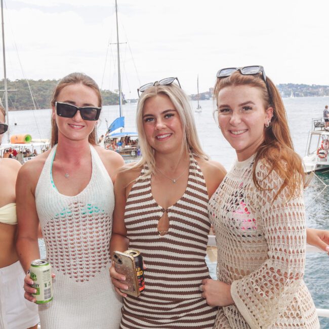 Four young women in summery knit outfits stand on a boat, smiling at the camera and holding drinks. Behind them, there are other boats and people on the water, suggesting a festive atmosphere.