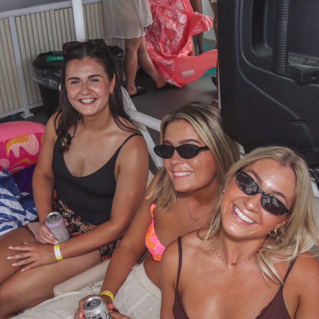 Three women in swimsuits and sunglasses smile at the camera while sitting together at a lively outdoor pool or boat party. People and floaties are visible in the background.