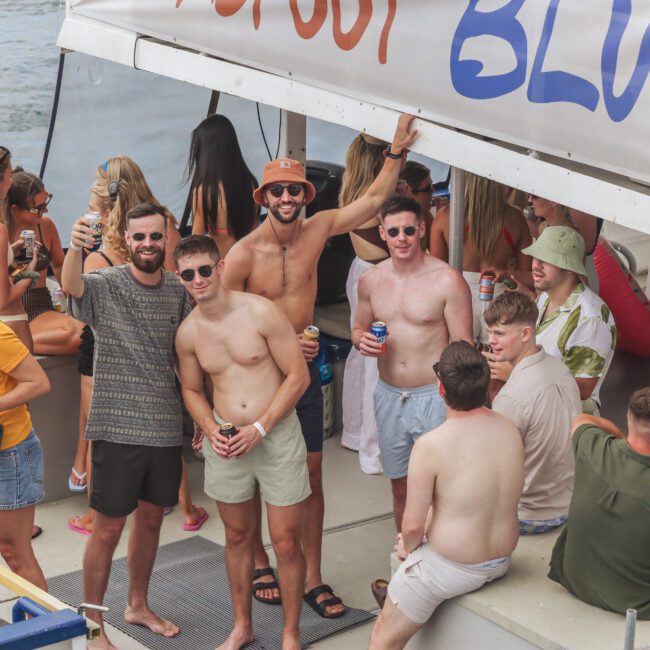 A group of young adults are partying on a boat, smiling and holding drinks. Some are wearing swimsuits and sunglasses. The atmosphere is lively, with people chatting and posing under a canopy labeled “BLUE” near the water.