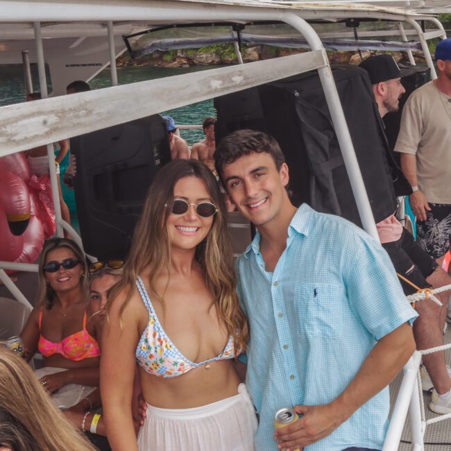 A smiling woman in a colorful bikini top and sunglasses stands next to a man in a blue shirt holding a drink, surrounded by people enjoying a party on a boat.