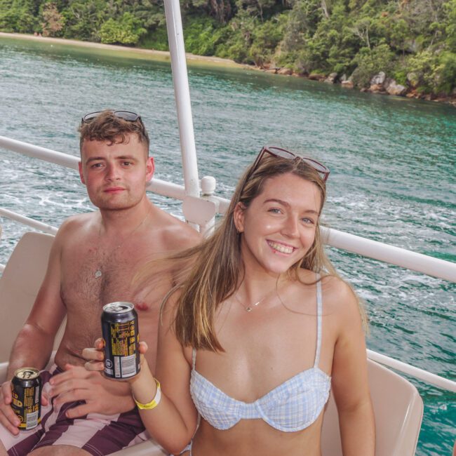 A young man and woman in swimsuits sit on a boat, smiling and holding canned drinks, with green water and a forested coastline visible in the background.