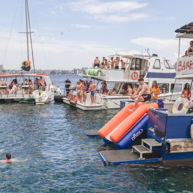 Several boats anchored close together host a lively party, with people swimming, sunbathing, and socializing. Some guests slide off a boat marked “Barefoot Blue” into the water, while others relax on inflatables.