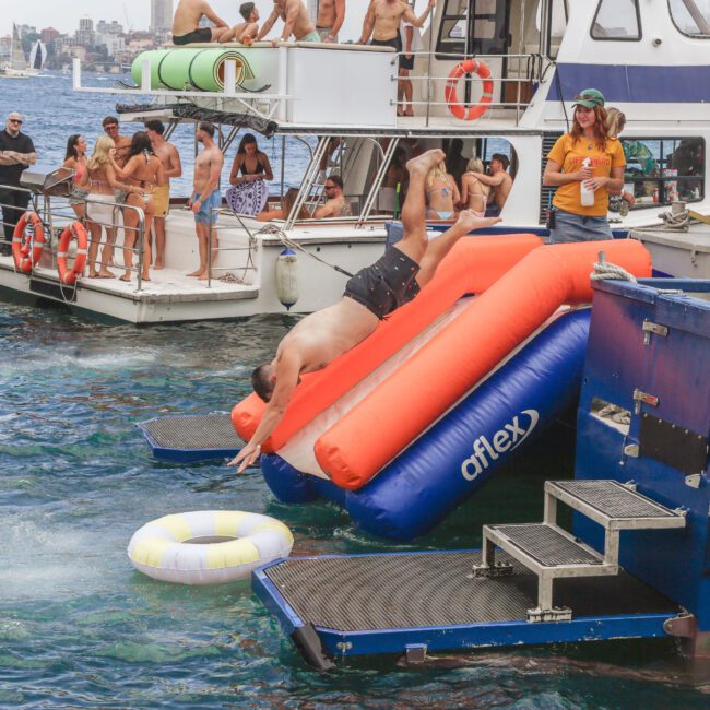 A man dives headfirst from an inflatable water slide into a lake, surrounded by boats and people in swimwear enjoying a sunny day. A few people watch and floatation rings are visible in the water.