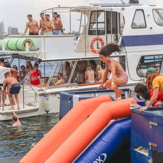A group of people enjoy a boat party; some are swimming, others relax on deck, while a woman slides down an orange slide into the water. The cityscape is visible in the background.