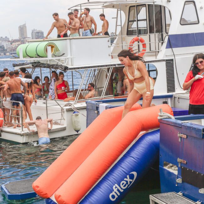 People enjoy a party on a boat. A woman in a bikini slides down an orange and blue inflatable slide into the sea, while others stand, swim, and relax on the deck and nearby. City buildings and water are visible in the background.