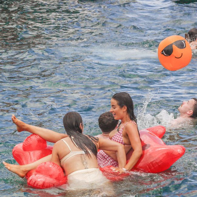 Three people sit on a red inflatable in a body of water, laughing. Nearby, one person floats on their back and another is on a pink float. An orange beach ball with sunglasses floats in the water.