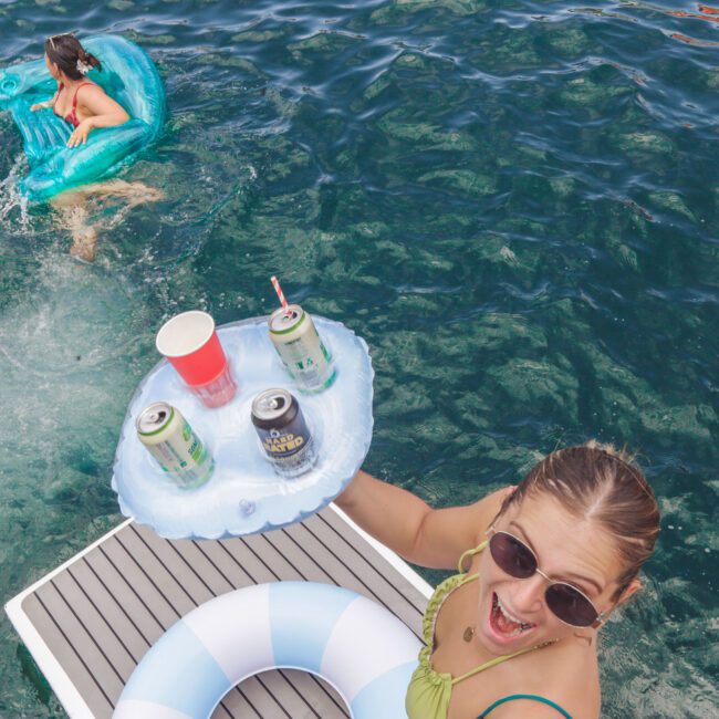 Two women enjoy a day in the water; one floats on an inflatable dolphin, while the other, wearing sunglasses and a swimsuit, holds a floating drink holder with cans and cups, smiling enthusiastically at the camera.