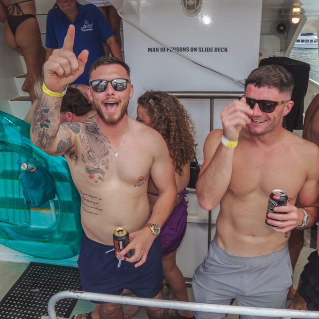 Two smiling men wearing sunglasses and swim shorts stand on a boat, holding drinks and gesturing enthusiastically. Other people in swimwear are around them, and an inflatable float and water are visible in the background.
