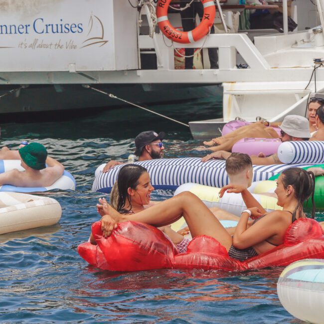 People relax on colorful inflatable loungers in the water near a boat labeled "Rum Runner Cruises." The scene is lively, with adults chatting and enjoying a sunny day on the water.
