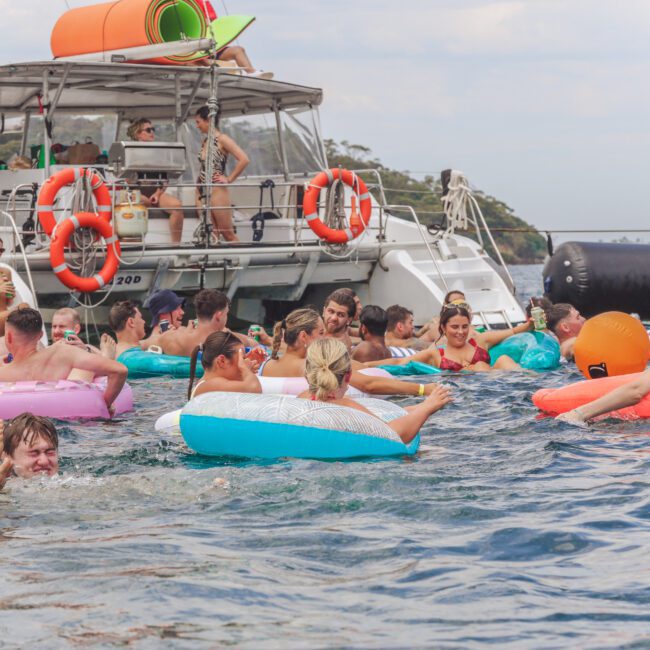 A group of people swim and relax on colorful inflatable tubes in the water near boats. Some people are on the boats, while others enjoy the water, socializing and having fun on a sunny day.
