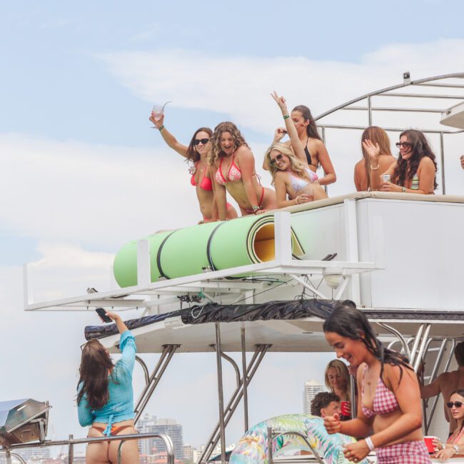 A group of women in swimsuits enjoy themselves on the upper deck of a yacht, waving and posing for photos, while others socialize and take pictures below on a sunny day. A man stands on the lower deck near a grill.