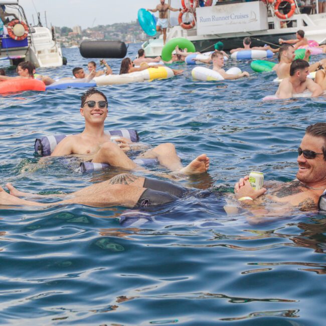 Two men relax on inflatable pool loungers in the water, smiling and enjoying drinks. Many people and colorful inflatables float nearby, with boats anchored in the background on a sunny day.