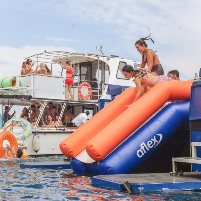People enjoy a summer day on the water with boats and an inflatable slide. Several individuals are sliding into the water while others watch and relax on the boats. The atmosphere is festive and playful.