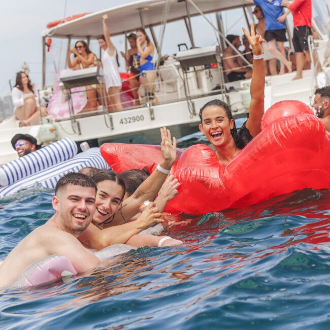 A group of young people smile and pose for the camera while swimming near a boat, holding colorful inflatables including a large red lips float. Other partygoers relax on the boat and in the water, enjoying a lively summer day.