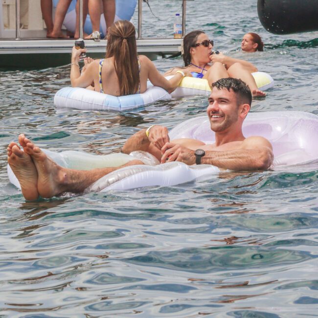 A man smiles while relaxing on a white inflatable float in a lake, surrounded by others on floats. Boats and people enjoying the water are visible in the background under a sunny sky.