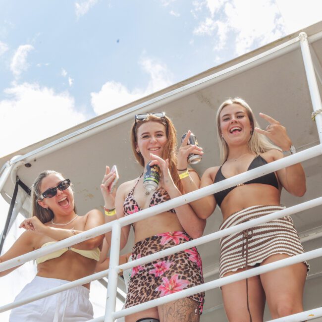 Three young women in swimsuits smile and pose for the camera, holding canned drinks on the deck of a white structure under a sunny, blue sky. They appear to be enjoying a summer outdoor event.