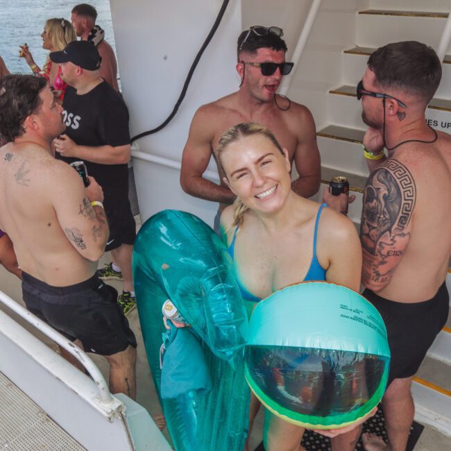 A group of people in swimwear are socializing on a boat. A smiling woman in the foreground holds an inflatable pool float and beach ball. Some people are drinking and standing by stairs. The mood is lively and relaxed.