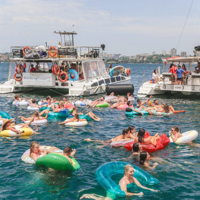 People relax on colorful inflatable tubes in the water near two catamaran boats on a sunny day. The boats are anchored close together, and the city skyline is visible in the background.