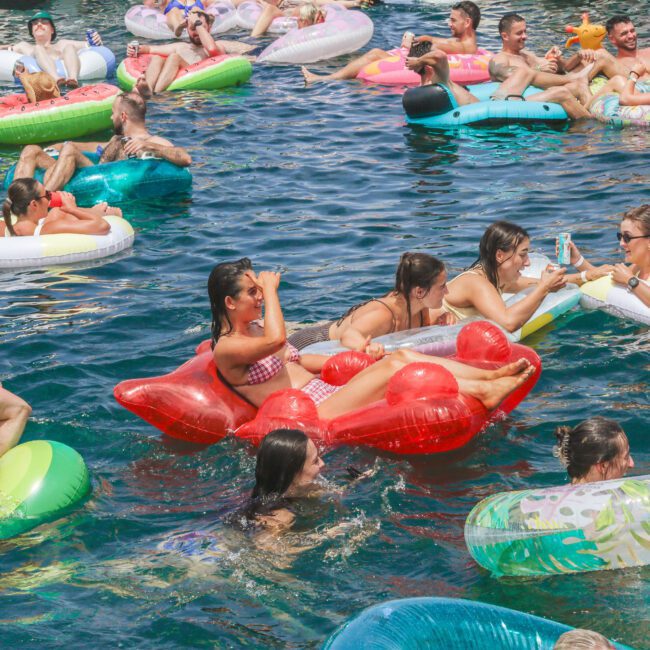 A group of people relax and socialize on colorful inflatable floats in a sunny, crowded pool or lake. Some are swimming, while others chat and laugh, enjoying the lively summer atmosphere.
