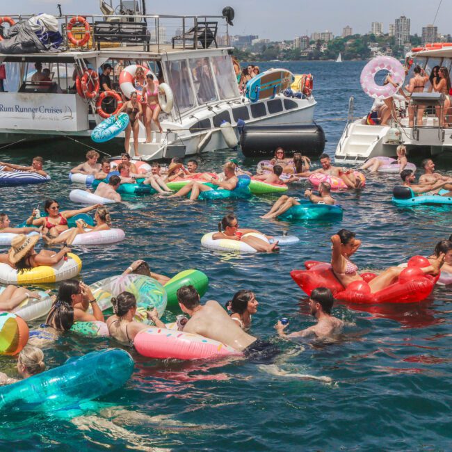 A group of people relax on colorful inflatables in the water near anchored boats on a sunny day. Some people are on the boats, while others float and chat in the lively scene. City buildings are visible in the background.