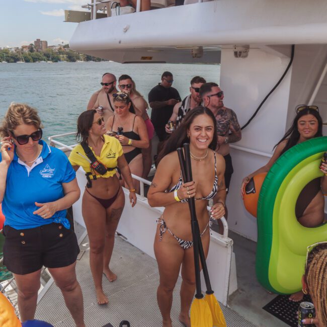 A group of people in swimsuits and casual clothing stand on a boat deck by the water. Some hold drinks, and one woman holds a paddle. An inflatable float is visible; everyone appears to be enjoying a sunny day.