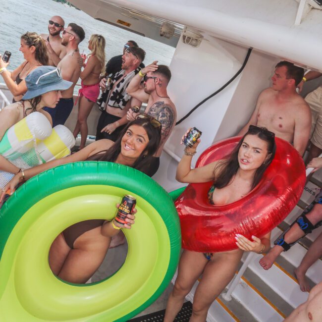 A group of people in swimsuits enjoy a boat party. Two women in front hold colorful inflatable rings and canned drinks, while others stand and socialize near the railing on a sunny day by the water.