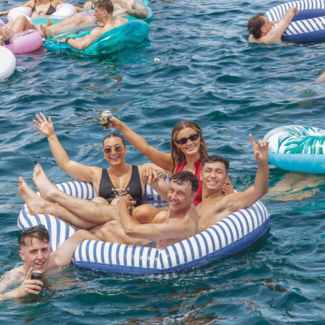 A group of young adults smile, pose, and hold drinks while lounging on striped and patterned inflatable rafts in clear blue water, surrounded by other people relaxing on floaties.