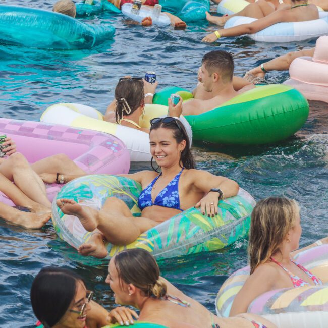 A group of people relax and socialize on colorful inflatable tubes in a body of water, enjoying a sunny day. Most are smiling and wearing swimsuits, creating a lively and cheerful summer scene.