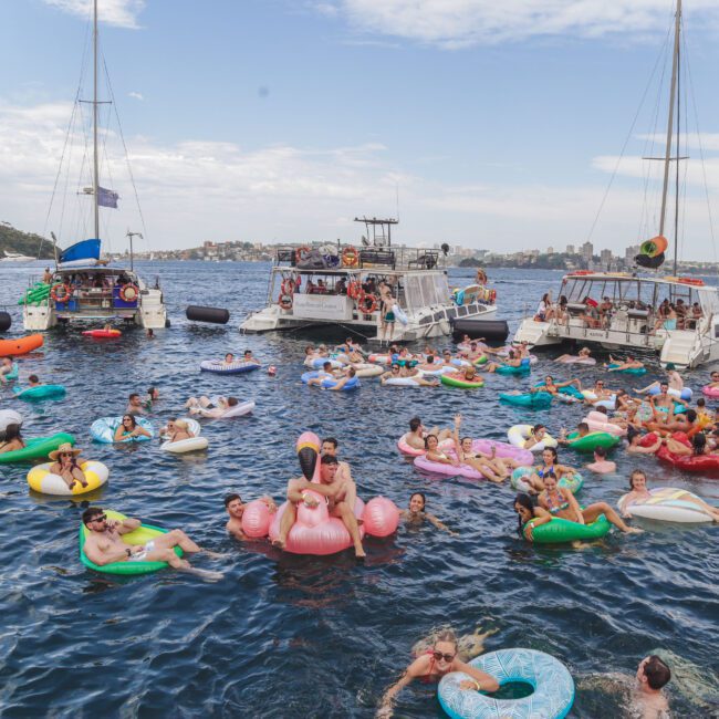A large group of people relax and have fun on colorful inflatable floats in the water near several anchored boats on a sunny day. The scene is lively and festive, with many people enjoying the summer atmosphere.