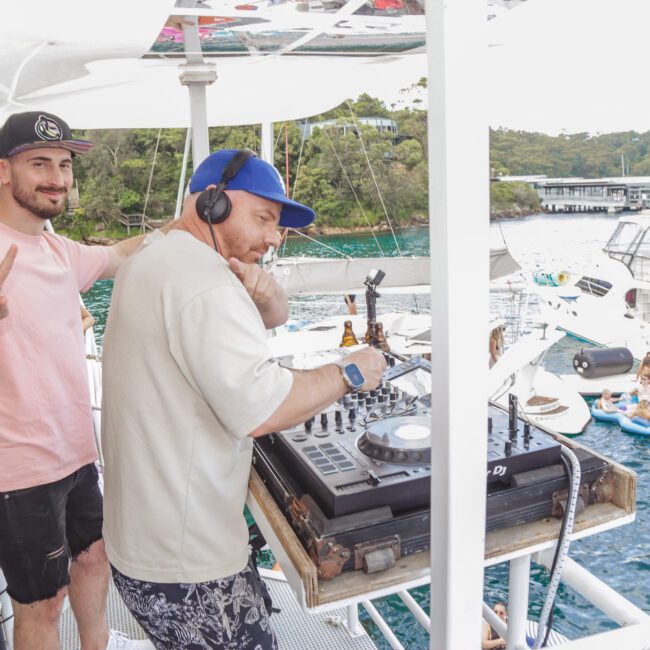 Two men on a boat, one is DJing with headphones while the other stands nearby flashing a peace sign. In the background, people relax on pool floats and boats on the water surrounded by trees.