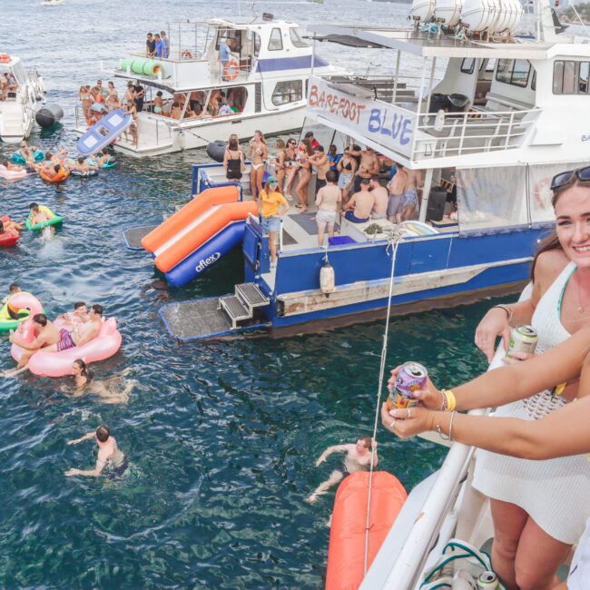 Two women smile at the camera on a boat, holding drinks. In the background, people swim and float on inflatables near several boats on a lively, sunny day on the water.
