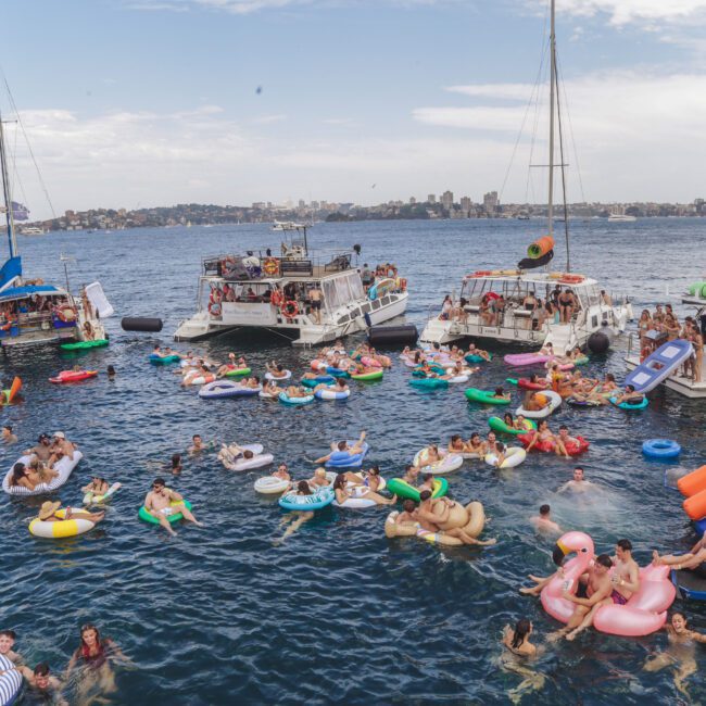 Dozens of people relax on colorful inflatables in the water between several anchored boats on a sunny day, with a city skyline and green hills visible in the background.