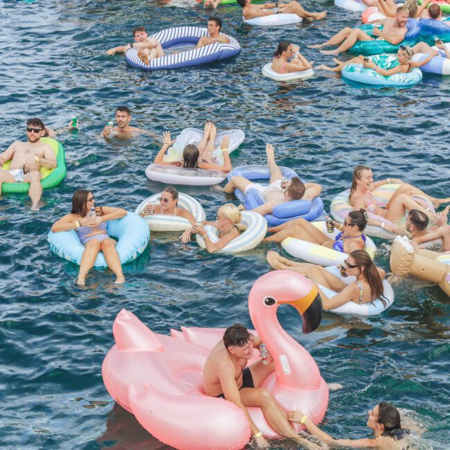 Groups of people relax on inflatable pool floats, including a large pink flamingo, in a body of water on a sunny day. Most are wearing swimwear and appear to be enjoying a festive atmosphere.
