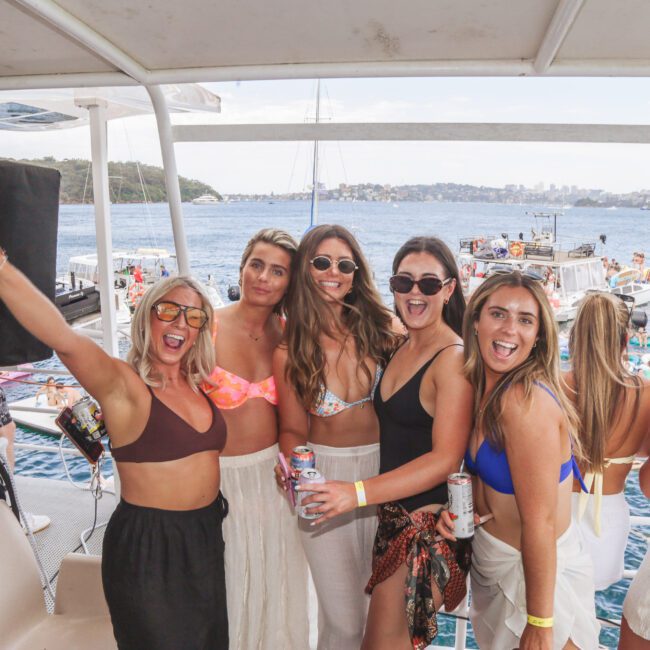 Five women in swimwear and sunglasses smile and pose for a group photo on a boat, holding drinks. Other people relax on boats in the water behind them on a sunny day.