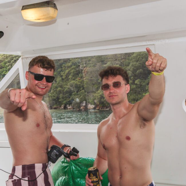 Two young men in swim trunks and sunglasses stand on a boat, smiling and pointing at the camera. One holds a drink and the other a speaker. Green water and forested shore are visible in the background.