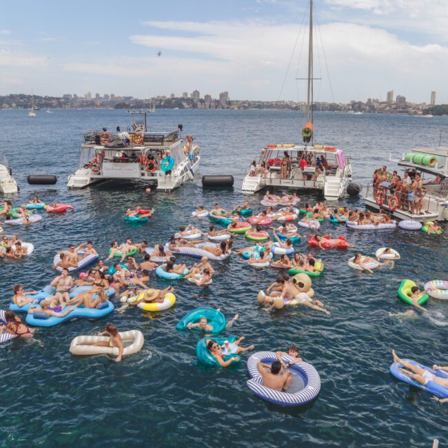 A large group of people relax on colorful inflatables in the water near several docked boats on a sunny day, with a city skyline visible in the background.