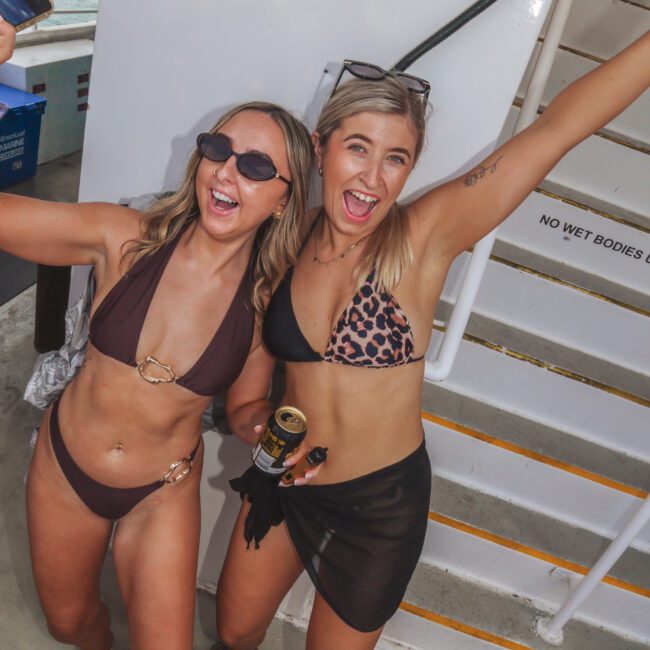 Two women in swimsuits smile and pose cheerfully on a boat deck, holding drinks. One wears a brown bikini, the other a leopard print top and black skirt. Water and railings are visible in the background.