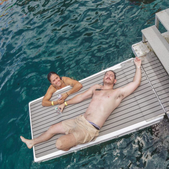 Two people relax on a floating dock by blue-green water; one is lying down in swim trunks holding a drink, and the other is sitting up, smiling at the camera near metal stairs leading into the water.