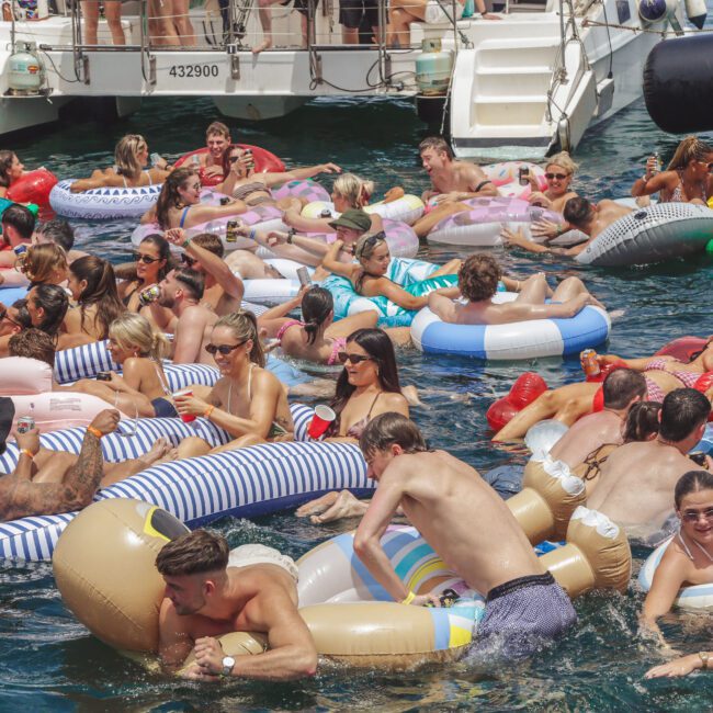 A large group of people relaxing on colorful inflatable pool floats in the water near a docked boat, enjoying a sunny day, with some people smiling, talking, and swimming.