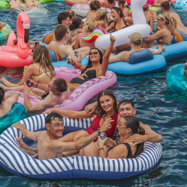 A crowded pool party with people relaxing on colorful inflatable floats, smiling, and holding drinks. Some floats are shaped like a flamingo and a unicorn. The atmosphere is lively and festive.