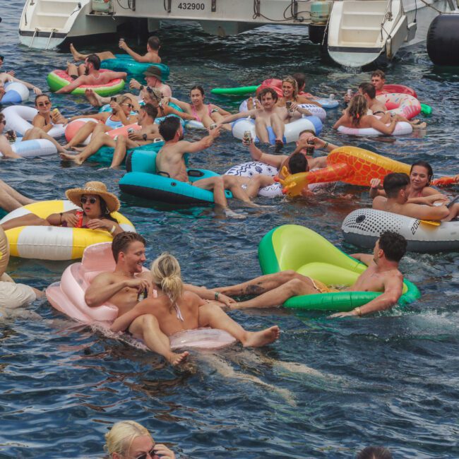 A large group of people float and relax on colorful inflatable tubes in the water near boats, enjoying a lively and crowded pool party atmosphere.
