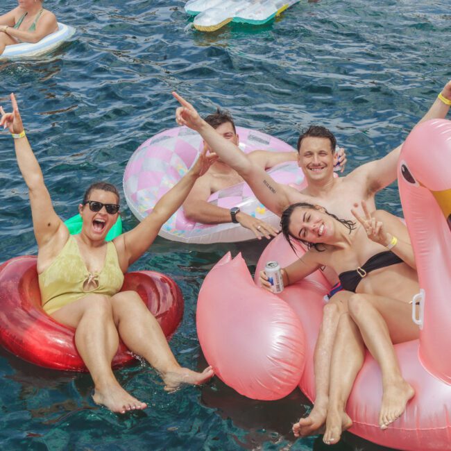 Four people smile and pose on colorful pool floats, including a pink flamingo, while holding drinks in a blue body of water surrounded by others on floats. They appear to be enjoying a lively summer day.