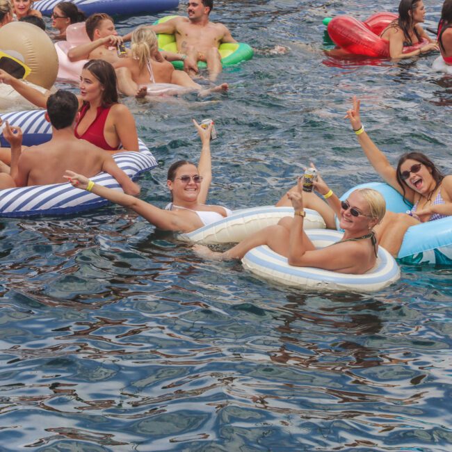 A group of people in swimsuits relax and have fun on colorful inflatable pool floats in the water, smiling and posing for the camera with drinks and peace signs during a lively outdoor gathering.