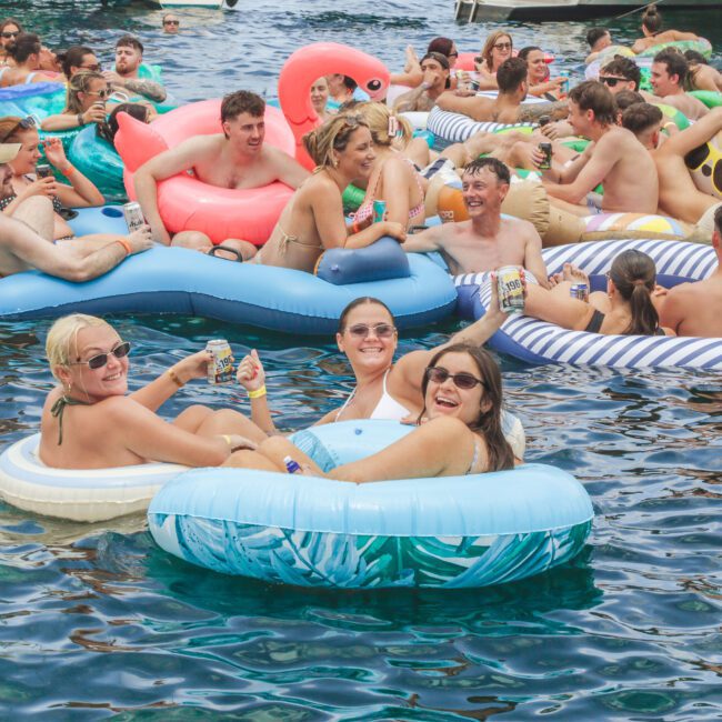 A large group of people relax on colorful inflatable floats in a body of water, socializing and enjoying drinks under sunny weather. Some floats are shaped like animals, creating a festive summer atmosphere.