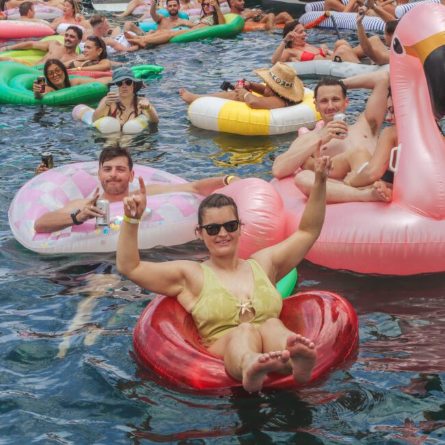 A group of people relax and have fun on colorful inflatable pool floats in the water near several boats, smiling, waving, and enjoying a lively summer party atmosphere.