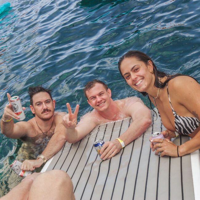 Three people in swimsuits smile and pose with drinks while sitting on a boat platform in clear blue water. One person flashes a peace sign. An inflatable float is visible in the background.