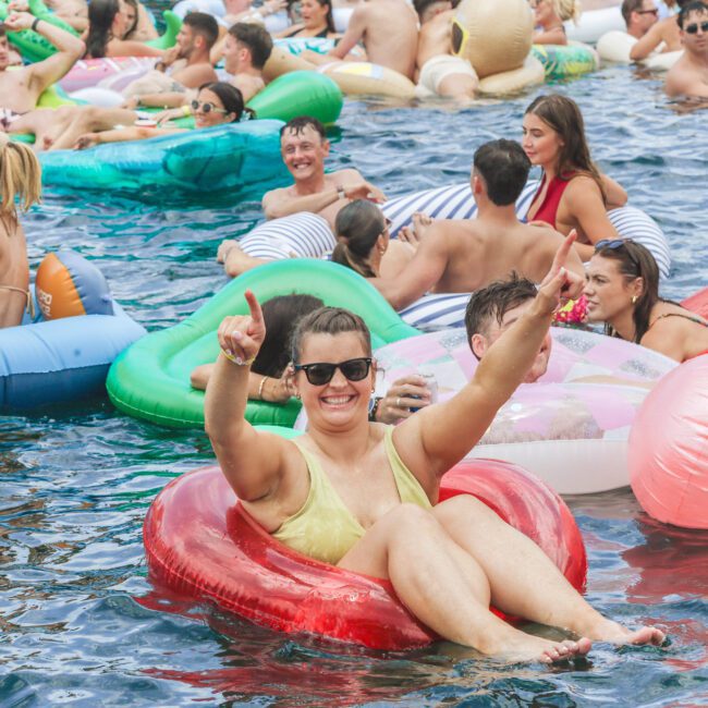 A woman in sunglasses and a yellow swimsuit smiles and raises her arms while lounging on a red pool float. Around her, many people relax on colorful inflatables in a crowded outdoor pool.