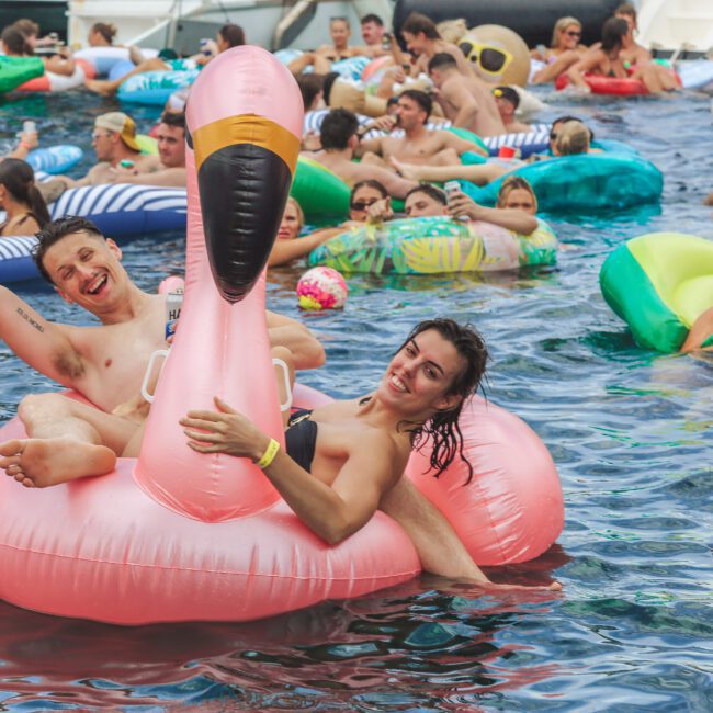 A large group of people enjoying a pool party, floating on colorful inflatables. Two people smile and pose on a giant pink flamingo float in the foreground, surrounded by others on various floaties.